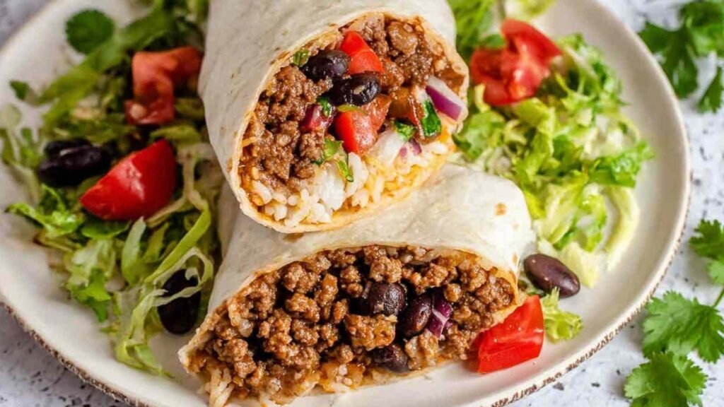 Beef Burrito Recipe 22 Homemade beef burrito recipe sliced in half showing seasoned ground beef, black beans, rice, and melted cheddar on a plate with salsa and sour cream