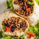 Beef Burrito Recipe 24 Homemade beef burrito recipe sliced in half showing seasoned ground beef, black beans, rice, and melted cheddar on a plate with salsa and sour cream