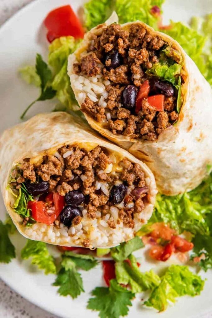 Beef Burrito Recipe 23 Homemade beef burrito recipe sliced in half showing seasoned ground beef, black beans, rice, and melted cheddar on a plate with salsa and sour cream