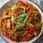 Chicken yakisoba in a large black skillet topped with sesame seeds and sliced green onions