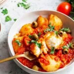 High Protein Bacalao a la Vizcaina served in a skillet with golden batter-fried salt cod, cannellini beans, potatoes, and green olives in tomato sauce