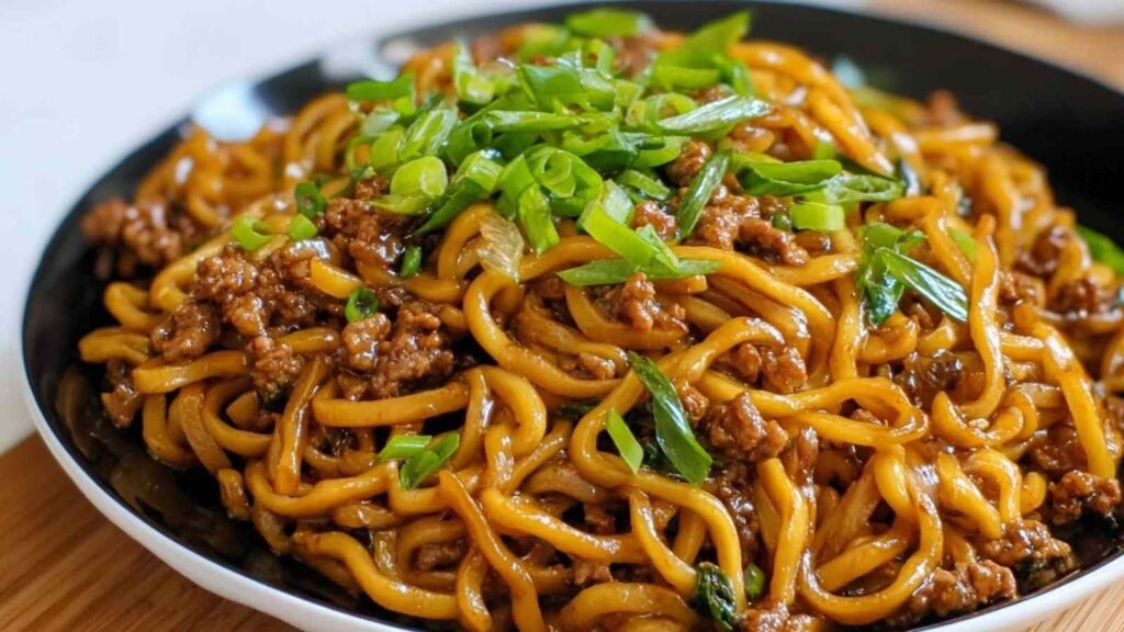 A bowl of high protein Mongolian noodles topped with sesame seeds and green onions on a dark background