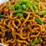 A bowl of high protein Mongolian noodles topped with sesame seeds and green onions on a dark background