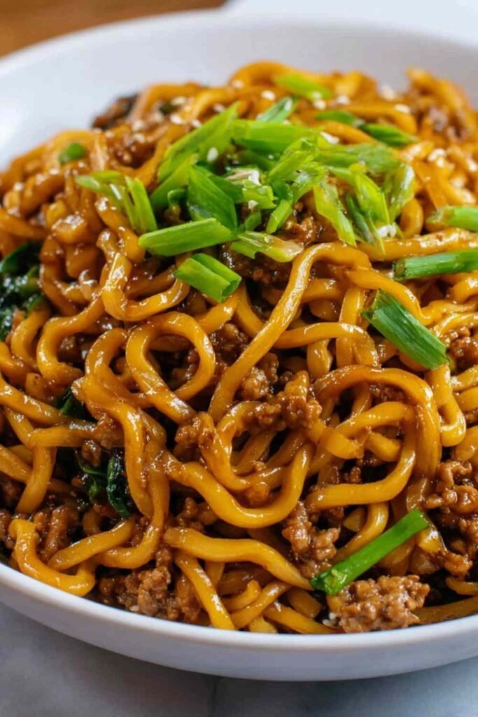 A bowl of high protein Mongolian noodles topped with sesame seeds and green onions on a dark background