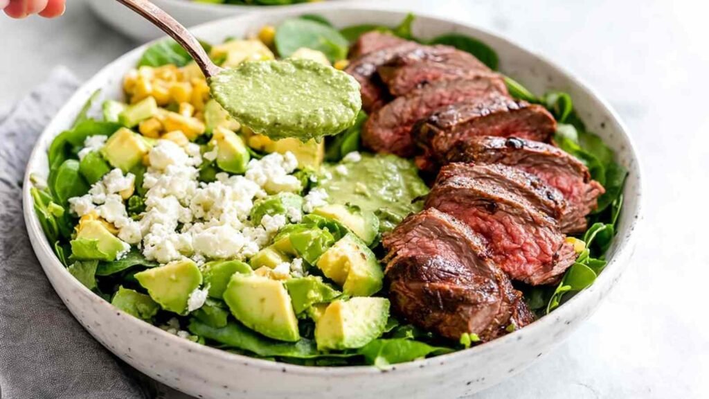 Steak avocado salad with grilled sirloin slices, diced avocado, corn, feta, and green goddess dressing in a white bowl