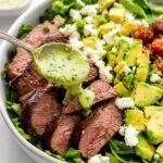 Steak avocado salad with grilled sirloin slices, diced avocado, corn, feta, and green goddess dressing in a white bowl