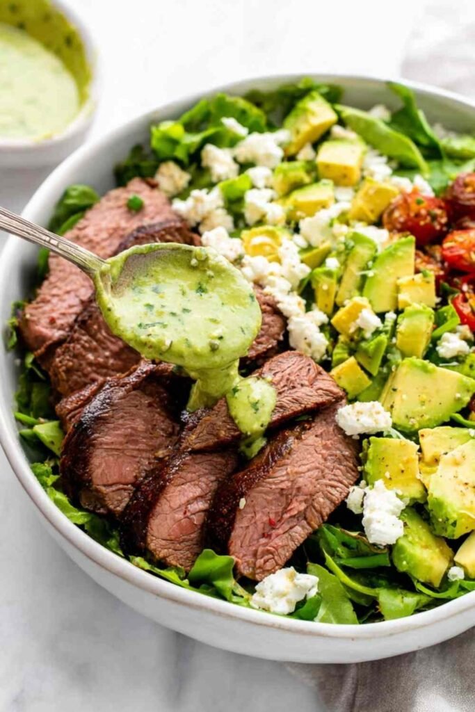 Steak avocado salad with grilled sirloin slices, diced avocado, corn, feta, and green goddess dressing in a white bowl