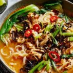 Overhead view of tahini gochujang noodle broth with turkey in a white bowl, topped with black sesame seeds, chili oil, spring onions, and lime wedge