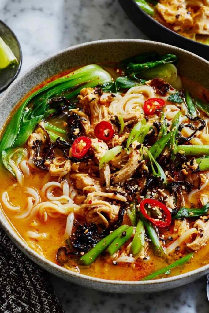 Overhead view of tahini gochujang noodle broth with turkey in a white bowl, topped with black sesame seeds, chili oil, spring onions, and lime wedge