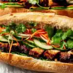 Vietnamese Chicken Banh Mi 18 Close-up of a Vietnamese Chicken Banh Mi on a wooden board with pickled carrots, fresh cilantro, jalapeno slices, and caramelized chicken on a toasted baguette