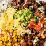 Overhead view of a DIY Chipotle steak bowl with grilled adobo steak, pico de gallo, corn salsa, guacamole, and Monterey Jack cheese over cilantro lime rice