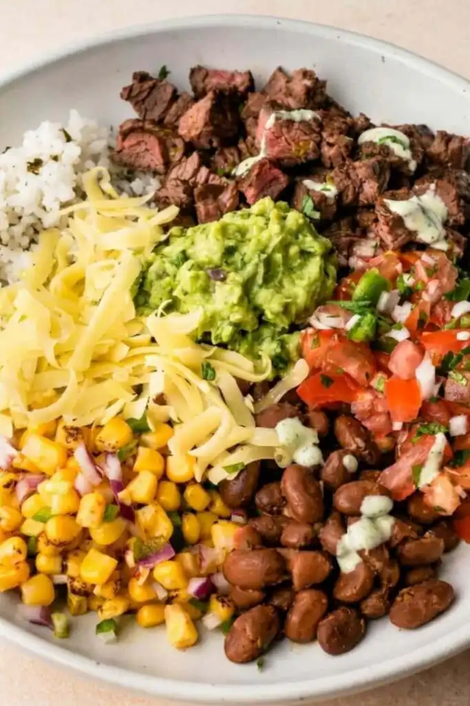 Overhead view of a DIY Chipotle steak bowl with grilled adobo steak, pico de gallo, corn salsa, guacamole, and Monterey Jack cheese over cilantro lime rice